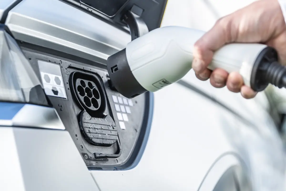 Une personne tient un connecteur de charge, s'apprêtant à le brancher au port de charge d'un véhicule électrique, des gouttelettes d'eau étant visibles.